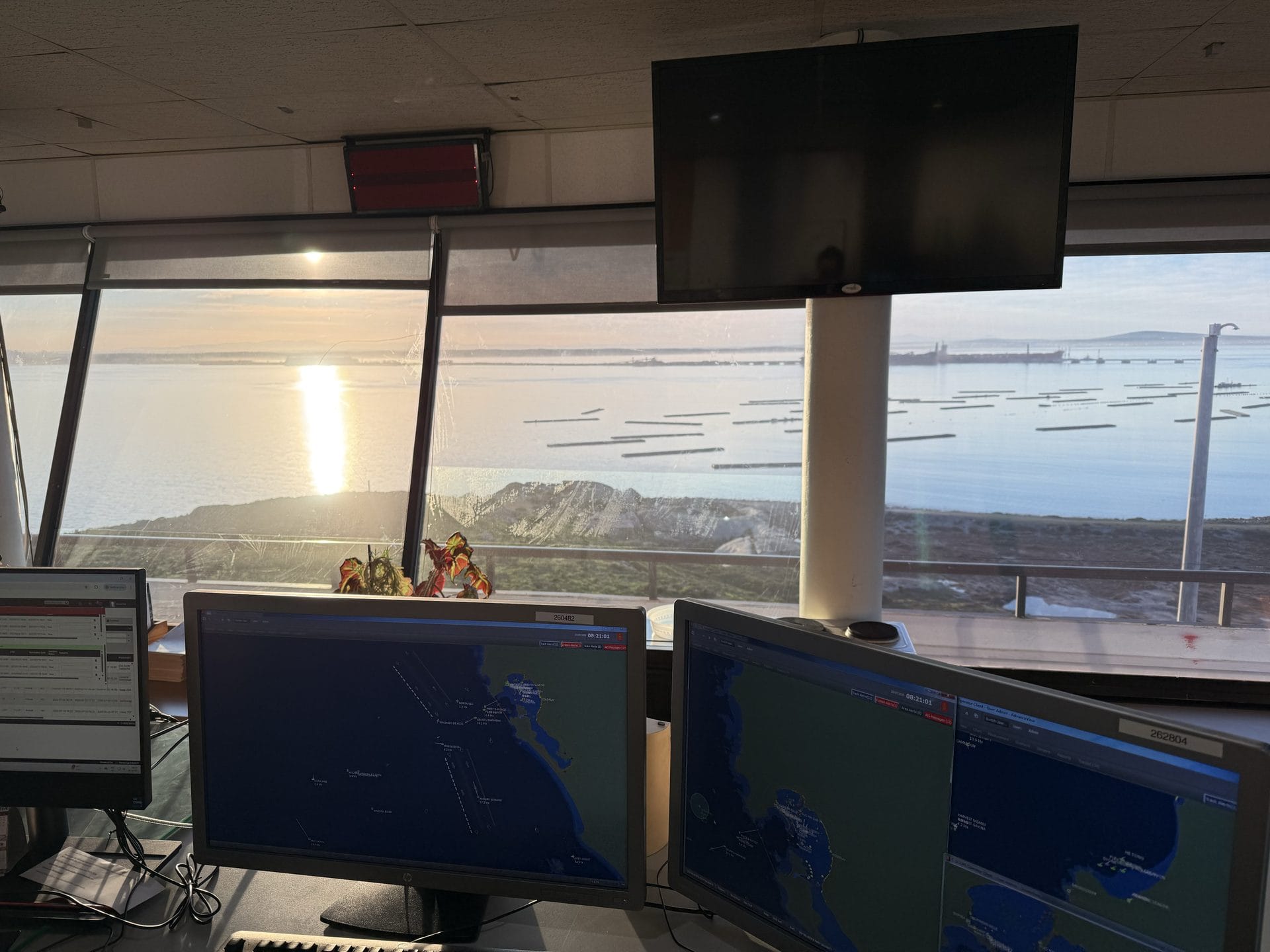 Operations vantage over Saldanha Bay, South Africa at last light: vessel-tracking and electronic-chart monitors with the bay, moored craft and headland beyond, where Afrishore coordinates west-coast port calls